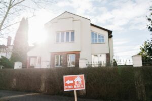 An orange "FOR SALE" sign with a white arrow and house icon stands in front of a modern, two-story cream-colored home under a bright, sunny sky.