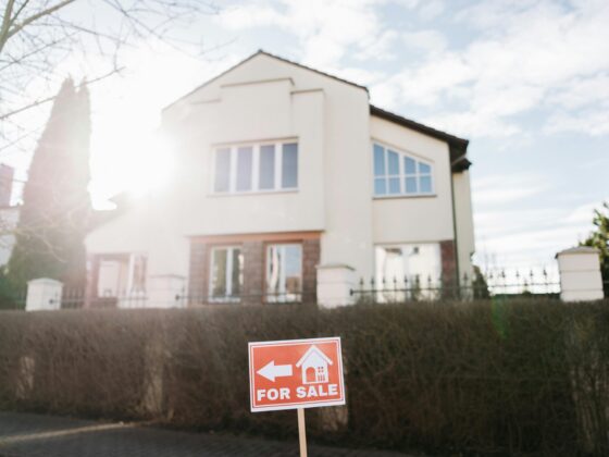 An orange "FOR SALE" sign with a white arrow and house icon stands in front of a modern, two-story cream-colored home under a bright, sunny sky.