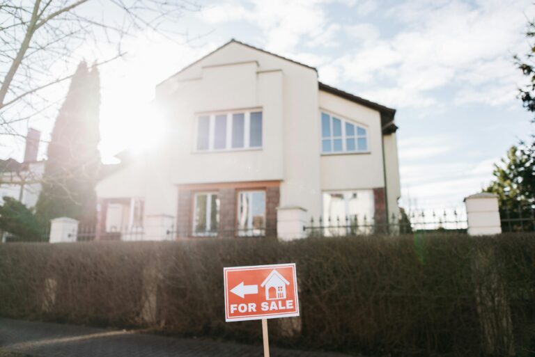 An orange "FOR SALE" sign with a white arrow and house icon stands in front of a modern, two-story cream-colored home under a bright, sunny sky.